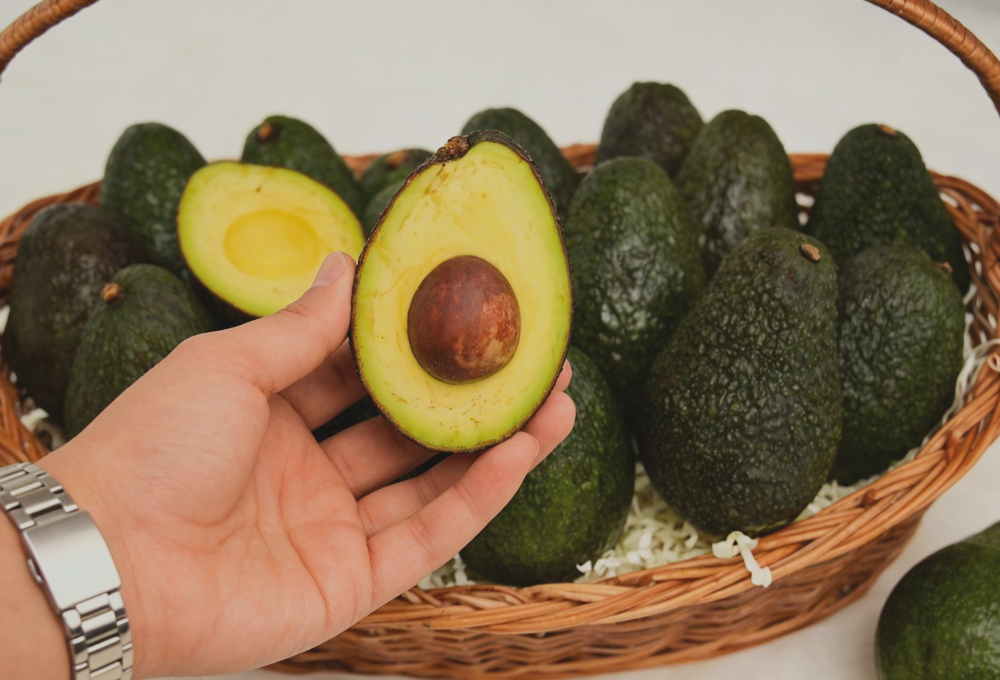 This image shows a hand holding a halved avocado, revealing its creamy flesh and seed, with a basket of whole avocados in the background.