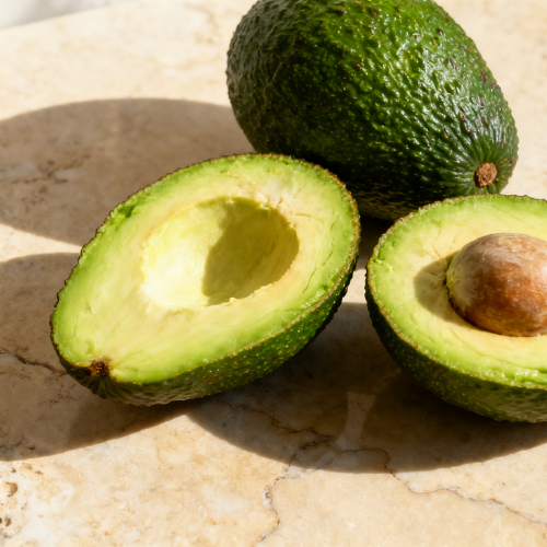 Whole and halved avocados with creamy green flesh are placed on a sunlit marble surface.