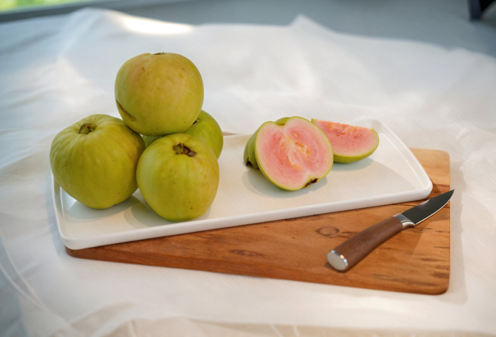 This image shows a pile of green guavas with one halved to reveal its pink interior, placed on a white platter and wooden cutting board, with a knife nearby.
