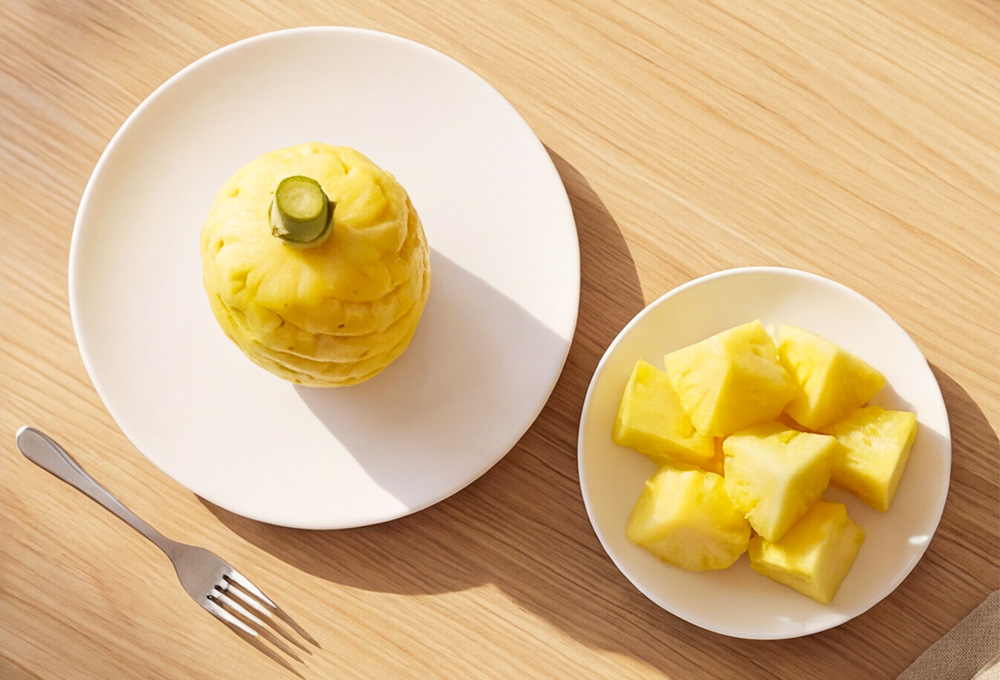 A fully peeled whole pineapple and fresh juicy pineapple chunks served on white plates atop a clean wooden surface with a fork.